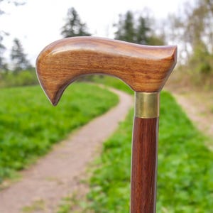 Puede incluir: Un bastón de madera con un mango curvo y un acento de latón. El bastón es marrón y tiene un acabado de madera natural.