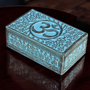 Puede incluir: Caja rectangular de madera con acabado turquesa y tallas florales intrincadas. La tapa presenta el símbolo Om en un medallón central. La caja es probablemente para guardar joyas u objetos pequeños.