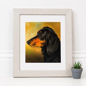 Puede incluir: Una lámina enmarcada que muestra un retrato de perfil de un perro salchicha negro y fuego. El perro aparece sobre un fondo cálido de color amarillo dorado. La obra está dentro de un marco de madera clara texturizada con un paspartú blanco.
