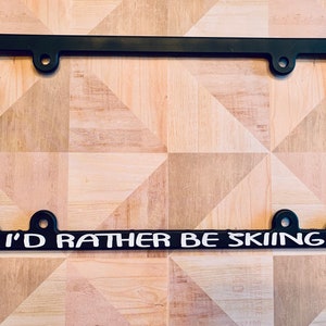 May include: Black plastic license plate frame with white text that reads "I'd rather be skiing" and a white ski goggle and hat graphic on each side.