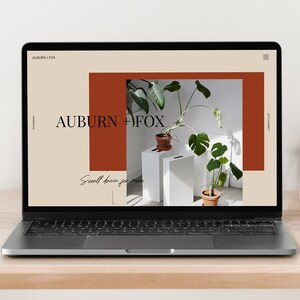 Op de afbeelding: Laptop met een website met de tekst "AUBURN + FOX". Het ontwerp gebruikt beige en roesttinten, met een plant en een klok op een houten bureau. De tekst "Scroll down for more" is ook zichtbaar.