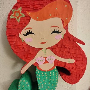 May include: A red and green mermaid piñata with gold glitter accents. The mermaid has red hair, a green tail, and a pink seashell bra.