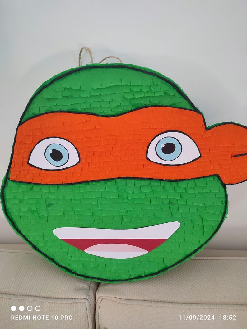 May include: Green and orange Teenage Mutant Ninja Turtles pi&ntilde;ata with a smiling face and blue eyes. The pi&ntilde;ata is made of paper mache and is shaped like a turtle's head.