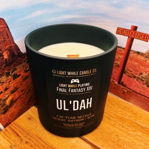 May include: A black candle in a matte finish jar with a white wax interior and a wooden wick. The label reads "Light While Playing: Final Fantasy XIV, Ul'Dah" with scent notes. The candle is on a wooden surface.