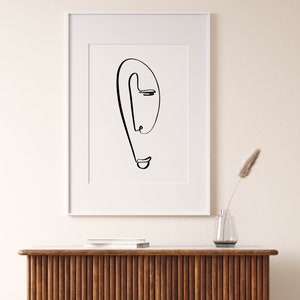 Puede incluir: Un dibujo en blanco y negro de una cara en un marco blanco. El dibujo es de una sola l&iacute;nea que forma el contorno de una cara, incluidos los ojos, la nariz y la boca. El dibujo est&aacute; sobre un fondo blanco y est&aacute; enmarcado en un marco blanco.
