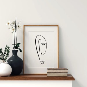 Puede incluir: Un dibujo en blanco y negro de un rostro con un solo ojo, enmarcado. El dibujo est&aacute; sobre un fondo blanco y el marco es de madera clara. El marco est&aacute; sobre una estanter&iacute;a de madera con dos libros apilados encima.