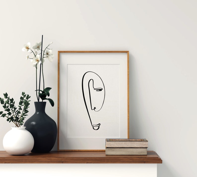Puede incluir: Un dibujo en blanco y negro de un rostro con un solo ojo, enmarcado. El dibujo est&aacute; sobre un fondo blanco y el marco es de madera clara. El marco est&aacute; sobre una estanter&iacute;a de madera con dos libros apilados encima.