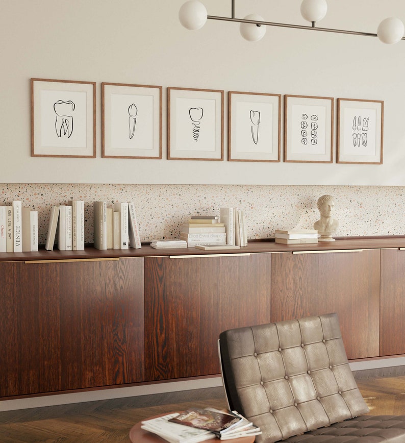以下が含まれることがあります： 白い壁に、自然な木のフレームに入った、歯のミニマルな白黒線画6枚が飾られています。その下には、金色の取っ手が付いたダークウッドのキャビネットがあります。