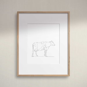 Puede incluir: Un dibujo en blanco y negro de una vaca en un estilo simple y minimalista. La vaca está representada de perfil, con la cabeza ligeramente girada hacia la derecha. El dibujo está enmarcado en un marco de madera claro.