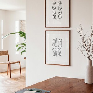 以下が含まれることがあります： 白い壁に、歯の白黒線画が額縁に入っています。上の絵は歯の格子状、下の絵は歯の列が描かれています。絵は、本と鉛筆が置かれた木製のテーブルの上に飾られています。