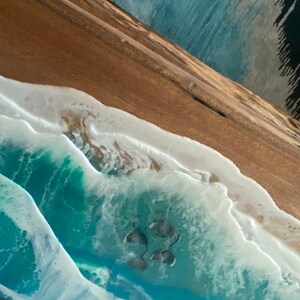 May include: A wooden table top with a resin ocean wave design. The wave is blue and green with white foam and a sandy bottom.