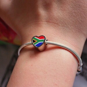 May include: A silver charm bracelet with a heart-shaped charm. The charm features the South African flag design in red, white, blue, yellow, and green. The bracelet is worn on a wrist.
