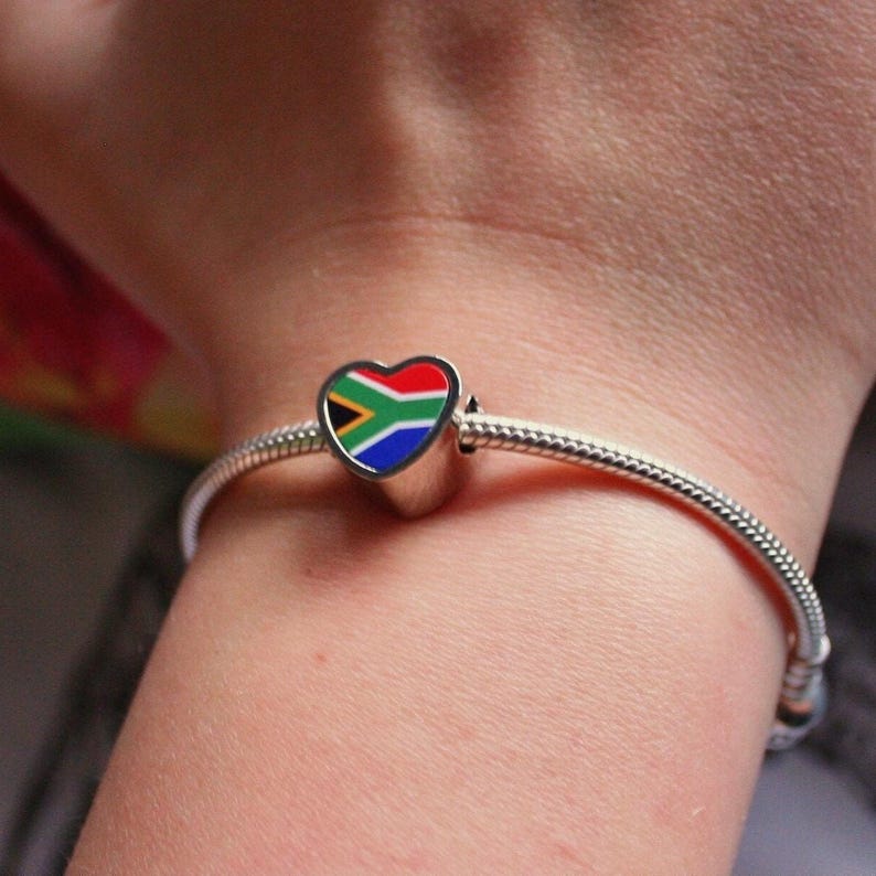 May include: A silver charm bracelet with a heart-shaped charm. The charm features the South African flag design in red, white, blue, yellow, and green. The bracelet is worn on a wrist.