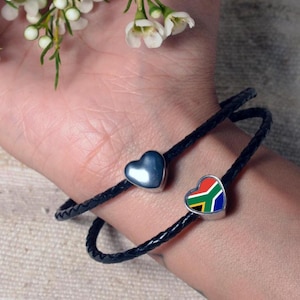 May include: Two black braided leather bracelets with heart-shaped charms. One charm is a solid silver heart, and the other features the South African flag. The bracelets are wrapped around a wrist.