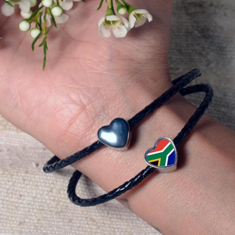 May include: Two black braided leather bracelets with heart-shaped charms. One charm is a solid silver heart, and the other features the South African flag. The bracelets are wrapped around a wrist.