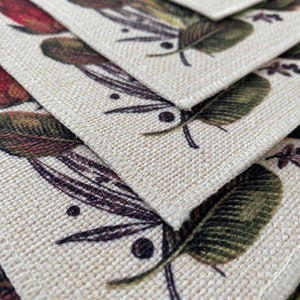 May include: Set of four beige linen placemats with a floral pattern featuring green leaves and red berries. The placemats have a stitched edge.