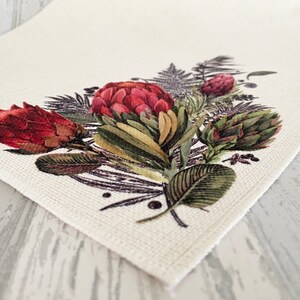 May include: A white fabric placemat with a floral design featuring red and green proteas, ferns, and other foliage.