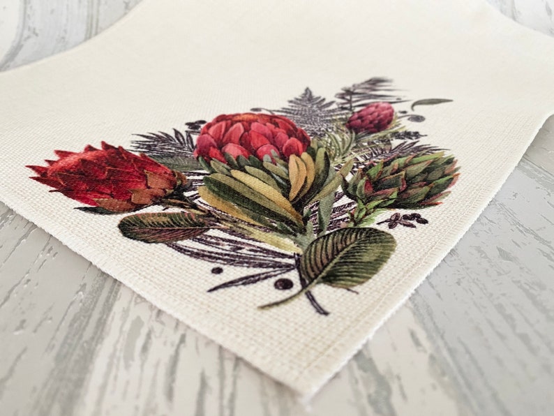 May include: A white fabric placemat with a floral design featuring red and green proteas, ferns, and other foliage.