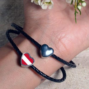 May include: A black leather bracelet with two heart-shaped charms. One charm is silver with a red and white striped design, and the other charm is silver and solid.