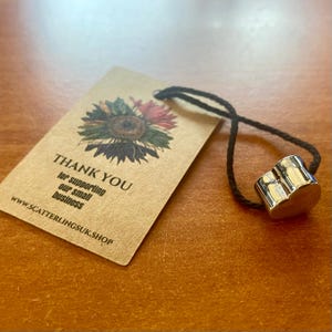 May include: A silver-colored, cylindrical bottle stopper with a black cord attached, next to a brown tag. The tag features a sunflower illustration and the words "THANK YOU" and "for supporting our small business". The website address is also visible.