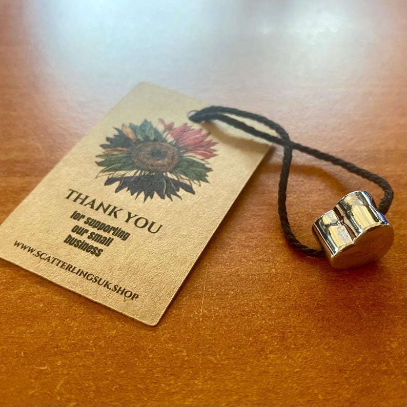 May include: A silver-colored, cylindrical bottle stopper with a black cord attached, next to a brown tag. The tag features a sunflower illustration and the words "THANK YOU" and "for supporting our small business". The website address is also visible.