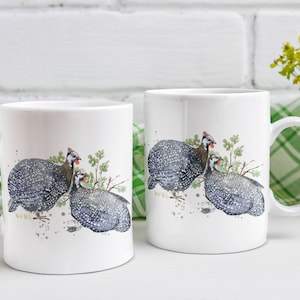 May include: Two white ceramic mugs with a black and white illustration of guinea fowl on a white background. The mugs have a black handle.