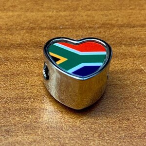 May include: A heart-shaped charm with a silver-colored metal frame. The charm features the South African flag design in red, white, blue, green, and yellow. The charm is likely for a bracelet or necklace.
