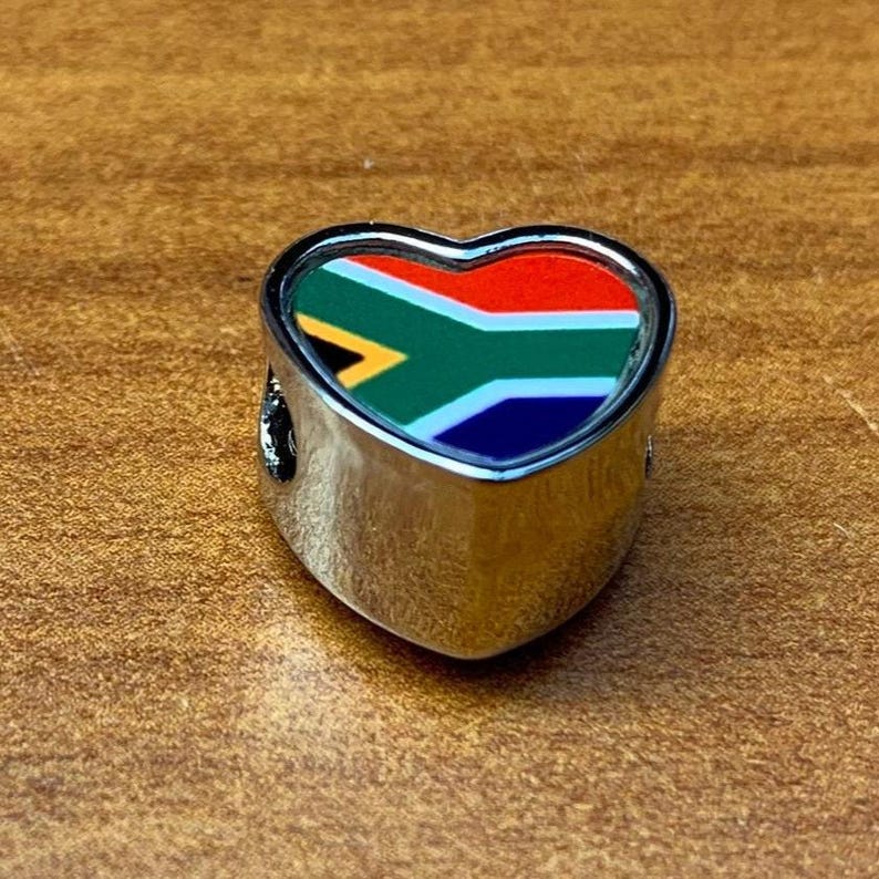 May include: A heart-shaped charm with a silver-colored metal frame. The charm features the South African flag design in red, white, blue, green, and yellow. The charm is likely for a bracelet or necklace.