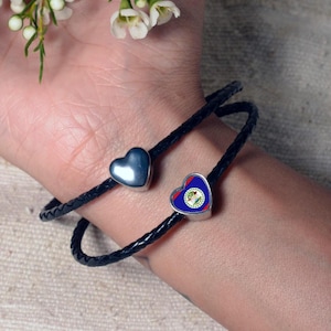 Könnte beinhalten: Ein schwarzes Lederarmband mit zwei herzförmigen Anhängern. Ein Anhänger ist silberfarben und der andere ist ein Herz mit der Flagge von Belize.