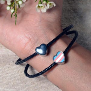 Könnte beinhalten: Ein schwarzes Lederarmband mit zwei herzförmigen Anhängern. Ein Anhänger ist silberfarben und der andere ist blau, rosa und weiß.