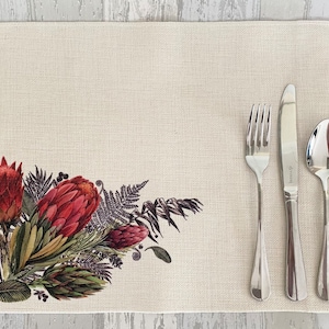 May include: A beige placemat with a floral design featuring red proteas, green leaves, and brown stems. The placemat has a textured surface and is set with a fork, knife, and spoon.