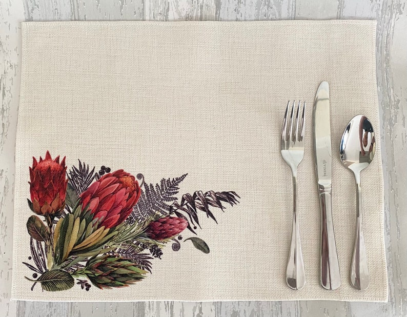 May include: A beige placemat with a floral design featuring red proteas, green leaves, and brown stems. The placemat has a textured surface and is set with a fork, knife, and spoon.