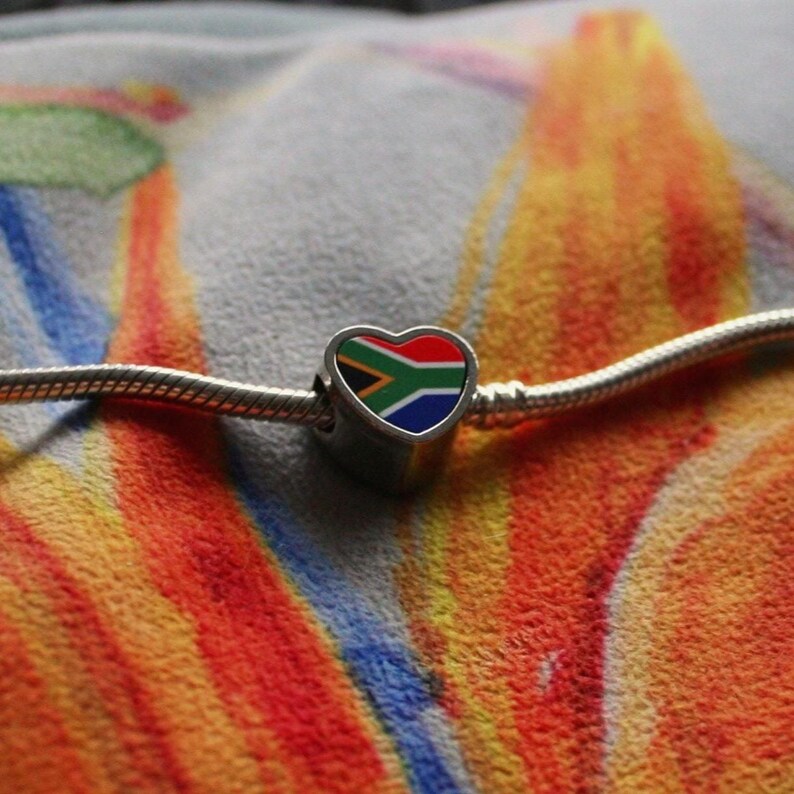 May include: A silver charm with a heart-shaped pendant featuring the South African flag design. The charm is on a silver chain, set against a colorful, textured background. The charm's design includes red, green, blue, yellow, and black colors.
