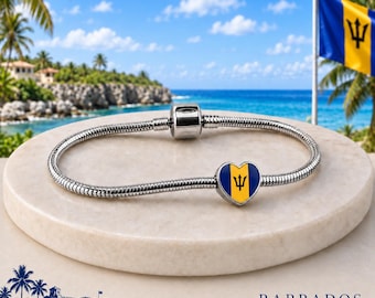 May include: A silver-toned snake chain bracelet featuring a heart-shaped charm. The charm displays the vertical blue, yellow, and blue stripes of the Barbados national flag with a black trident symbol centered on the yellow band.