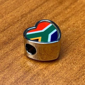 May include: A heart-shaped charm with the South African flag design. The charm features a silver-colored metal frame and a colorful enamel inlay of the flag's red, green, blue, yellow, and white elements. The charm has a hole for threading.