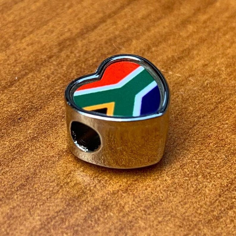 May include: A heart-shaped charm with the South African flag design. The charm features a silver-colored metal frame and a colorful enamel inlay of the flag's red, green, blue, yellow, and white elements. The charm has a hole for threading.