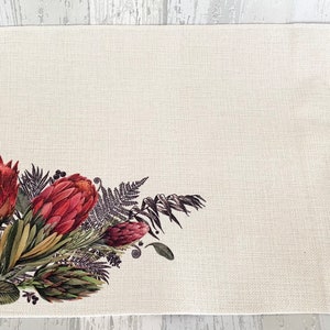 May include: A white placemat with a floral design featuring red proteas and green foliage. The design is in the bottom left corner of the placemat.