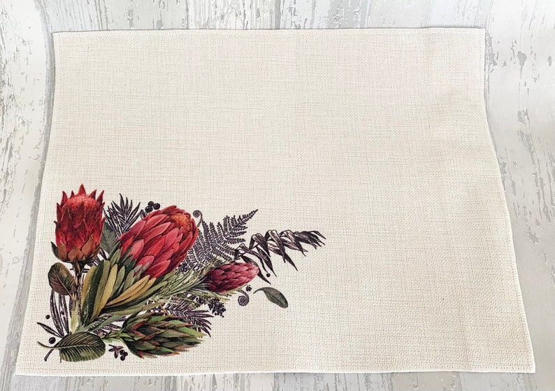 May include: A white placemat with a floral design featuring red proteas and green foliage. The design is in the bottom left corner of the placemat.