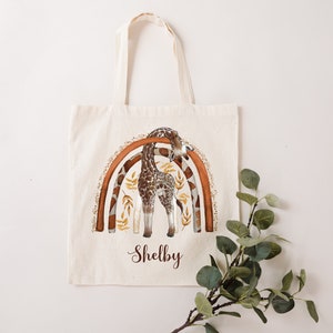 May include: A beige canvas tote bag with a brown and orange rainbow design featuring a giraffe. The bag has the name "Shelby" printed on the front.