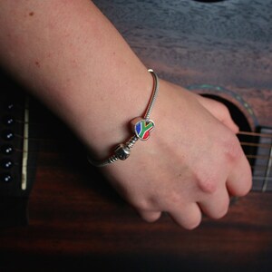 May include: A silver charm bracelet with a colorful charm featuring the South African flag design. The charm is heart-shaped with red, blue, green, and yellow accents. The bracelet is worn on a wrist, with a guitar in the background.