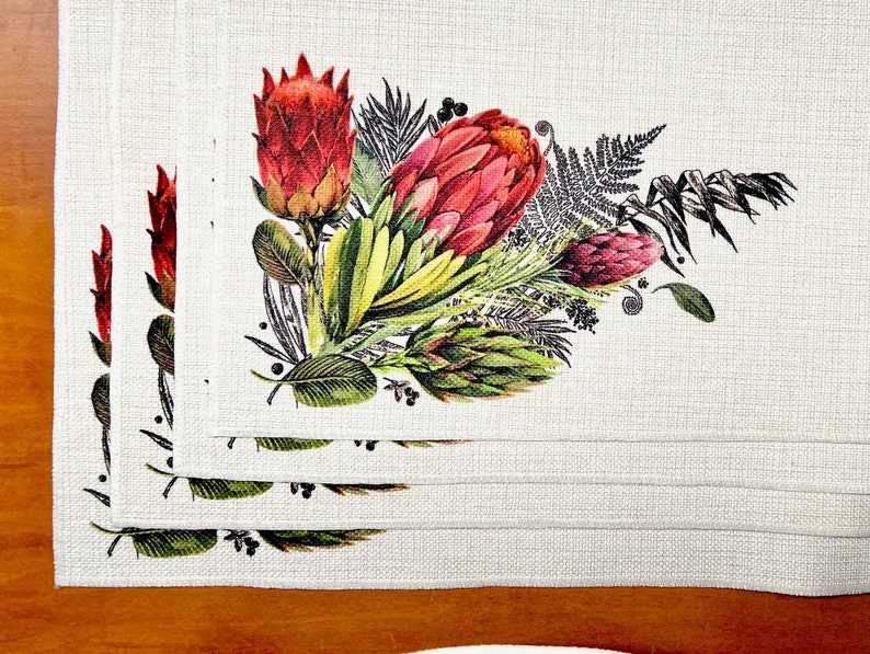 May include: Set of four white linen placemats featuring a floral design with red and green proteas, ferns, and other foliage.