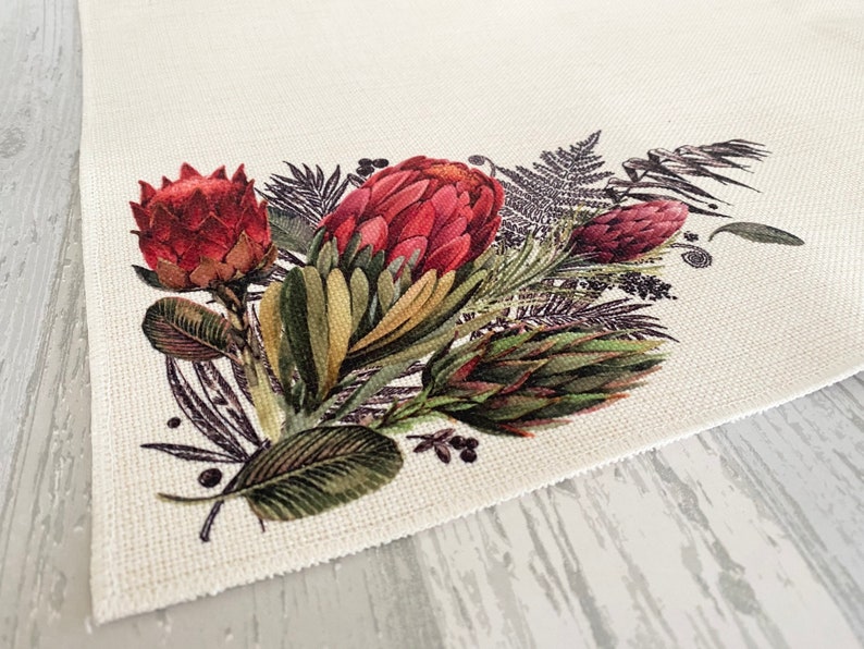 May include: A white fabric placemat featuring a printed design of red and green protea flowers and leaves.