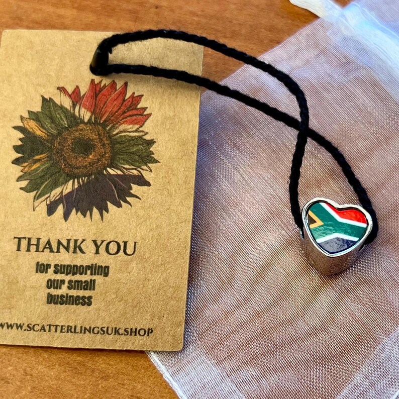 May include: A silver heart-shaped pendant with the South African flag design, suspended on a black cord. A brown tag with a sunflower illustration and the text "THANK YOU for supporting our small business" is visible.