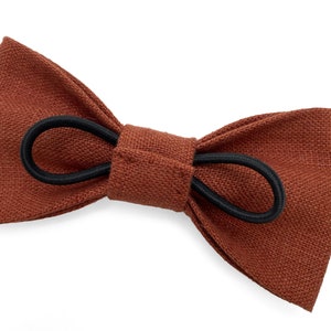 May include: A brown bow tie with a textured fabric appearance. The bow tie is secured with a black elastic band. The accessory is designed to be worn around the neck and is suitable for formal or casual wear.