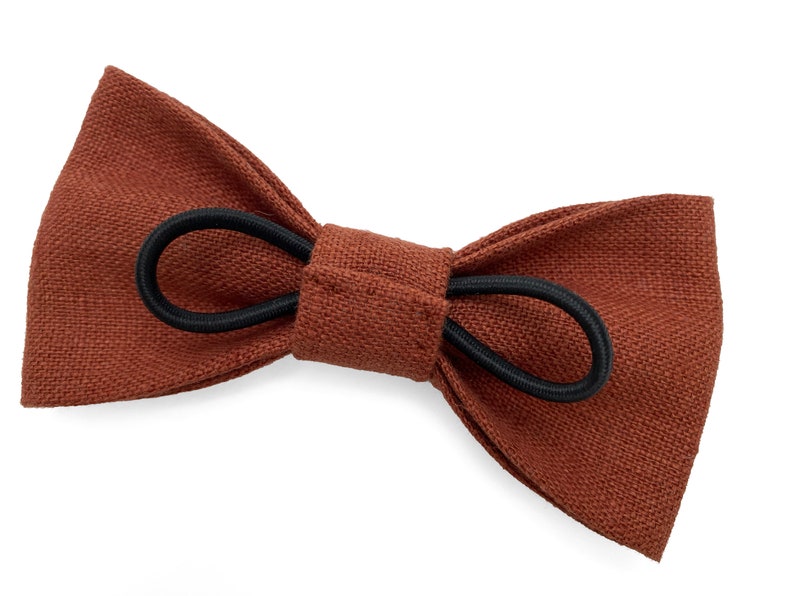 May include: A brown bow tie with a textured fabric appearance. The bow tie is secured with a black elastic band. The accessory is designed to be worn around the neck and is suitable for formal or casual wear.