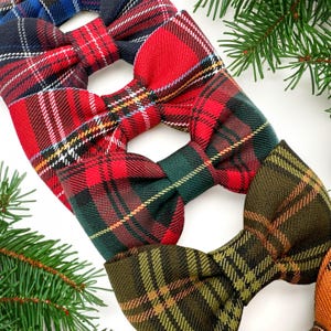 May include: A collection of plaid bow ties in various colors, including red, green, and blue. The bow ties feature classic tartan patterns with contrasting stripes. The image is set against a white background with evergreen sprigs.