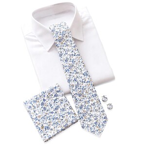 Peut inclure: Une chemise habillée blanche accompagnée d'une cravate, d'une pochette assortie et d'une paire de boutons de manchette ronds. La cravate et la pochette présentent un motif floral dense dans des tons bleu clair, marine et beige sur fond blanc.