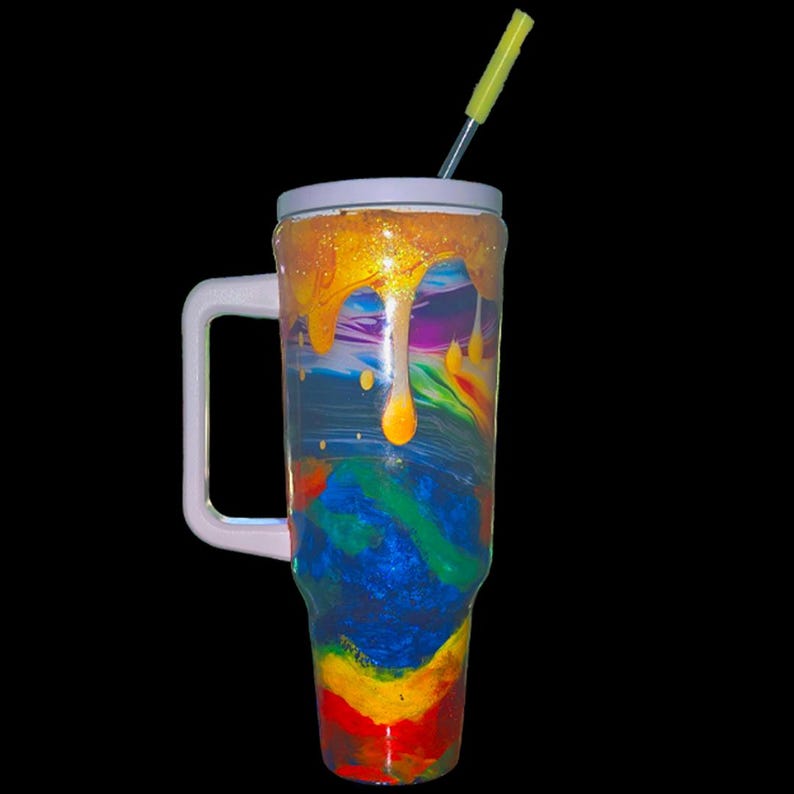 May include: A tall, cylindrical tumbler with a white handle and lid, featuring a vibrant, multi-colored swirl pattern of blue, green, yellow, and red. The top section has a glossy, dripping gold effect, and a yellow straw is inserted into the lid.
