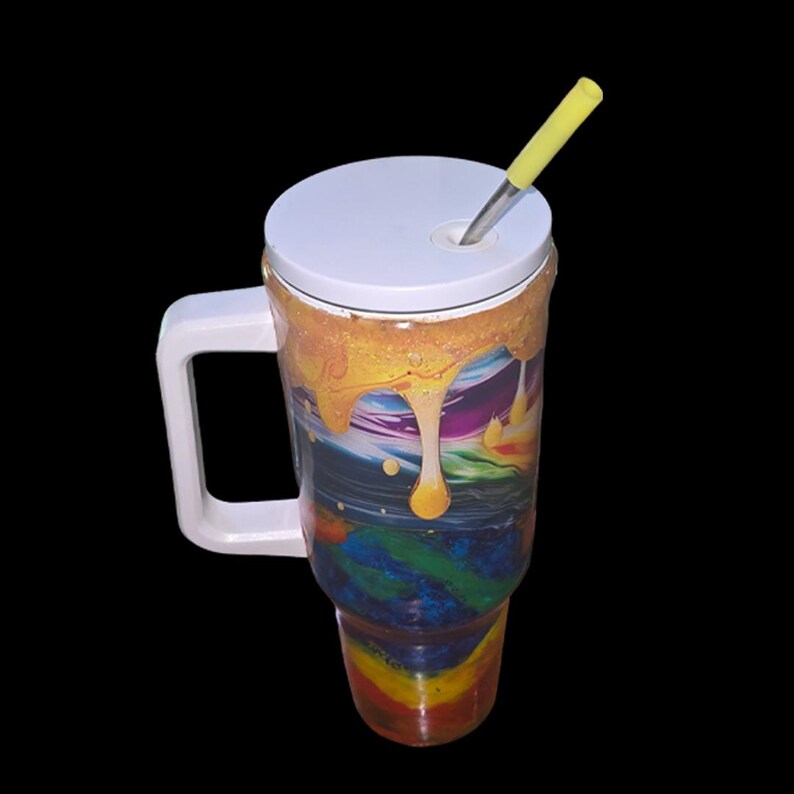 May include: A tall tumbler with a white handle and a white lid featuring a yellow straw. The exterior surface displays a swirling, multi-colored pattern of blue, green, and red, topped with a textured, dripping golden-yellow resin effect.