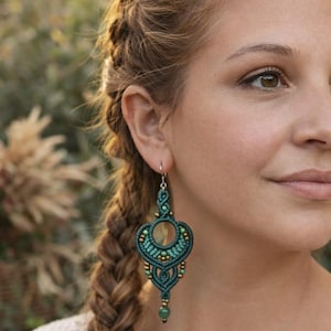 May include: A teal macrame earring with a circular design and small gold beads. The earring has a teardrop shape with a green stone at the bottom. The woman's hair is in a braid.